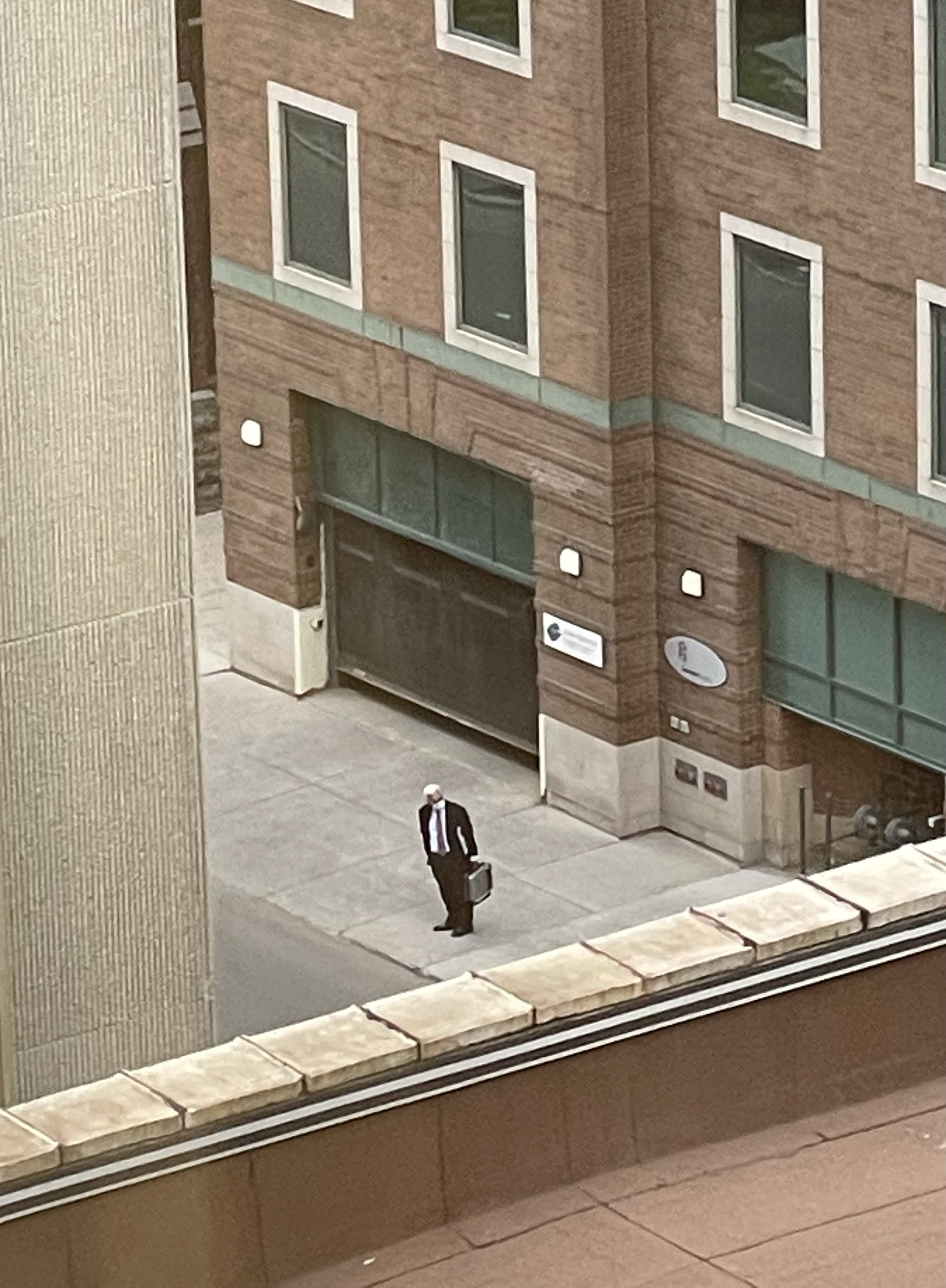 A man in a suit holding a briefcase stands alone on a wide empty sidewalk. A large brick building rises behind him. He’s not moving. He is looking at something outside the frame.